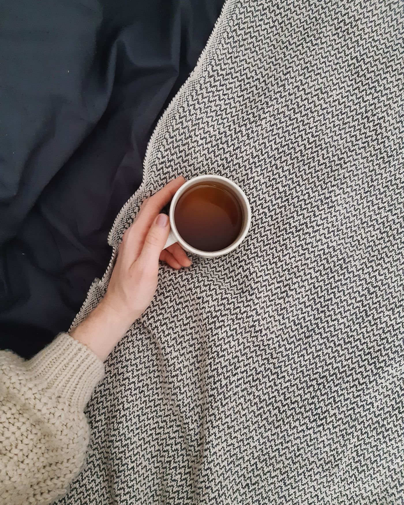 A hand holding a warm cup of tea beside a blanket